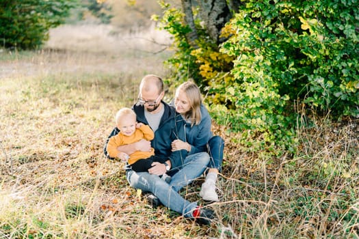 Mom hugs dad with a little boy in his arms sitting in a clearing in the park. High quality photo