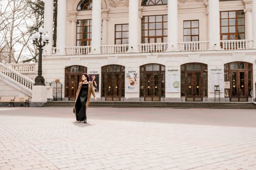 Woman street lifestyle. Image of stylish woman walking through European city on sunny day. Pretty woman with dark flowing hair, dressed in a beige raincoat and black, walks along the building