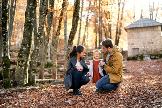 Dad and mom are squatting and looking at a little squinting girl in the autumn forest. High quality photo