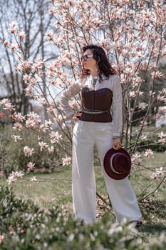 Magnolia park woman. Stylish woman in a hat stands near the magnolia bush in the park. Dressed in white corset pants and posing for the camera