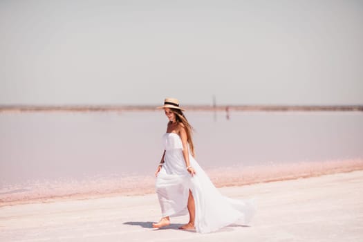 Woman in pink salt lake. She in a white dress and hat enjoys the scenic view of a pink salt lake as she walks along the white, salty shore, creating a lasting memory