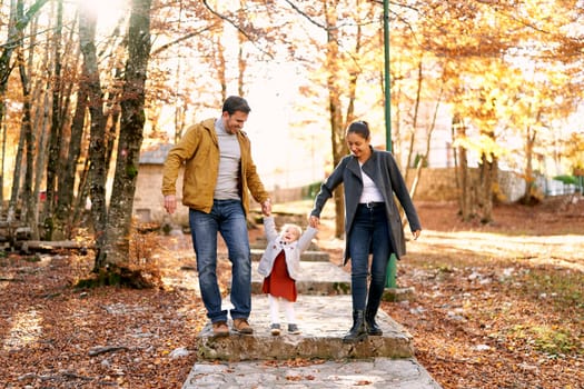 Little girl stands on a paved path in the park holding hands with mom and dad. High quality photo