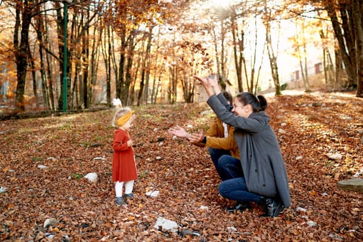 Little girl stands in front of mom and dad squatting and throwing up leaves in the autumn park. High quality photo