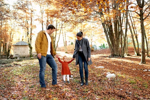 Mom and dad look at a little girl, holding her hands in the autumn forest. High quality photo