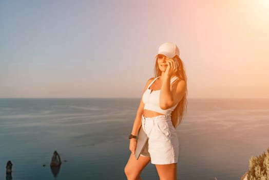 A happy woman in white shorts and T-shirt enjoys the picturesque sea view while holding a laptop and talking on the phone