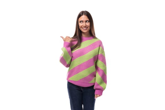 stylish cheerful young brunette woman in a striped pink sweater points her finger to the side on a white background with copy space.