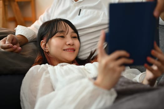 Pretty young Asian woman lying on couch with husband and reading interesting book.