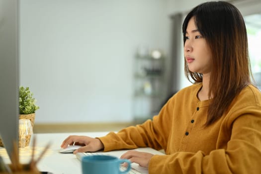 Concentrate young Asian female looking at computer screen working online at home.