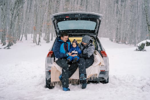 Mom and dad with a small child are sitting in the trunk of a car under a snowfall. High quality photo