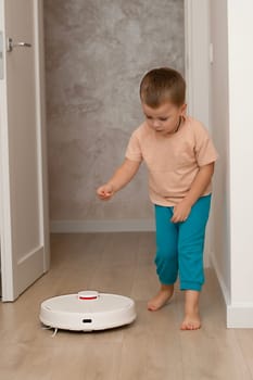 Cleaning concept. A little funny Caucasian boy, 4 years old, throws torn paper in small pieces onto the floor for cleaning and suction by a white robot vacuum cleaner. Plays happily in the home interior.