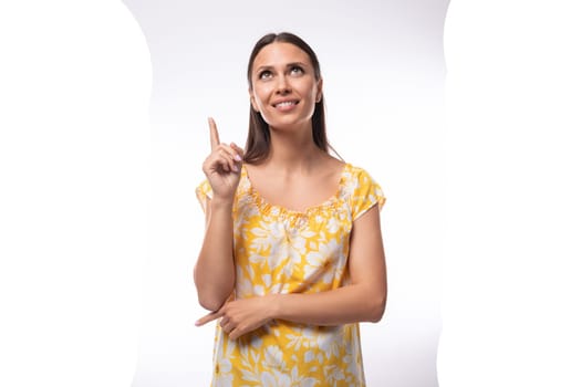 Caucasian young woman with straight black hair dressed in a yellow sundress gesturing with her fingers.