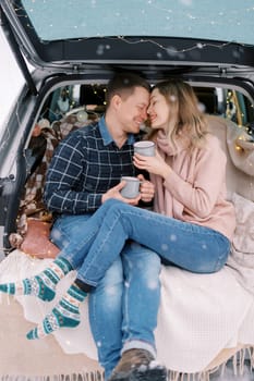 Smiling husband and wife sitting with cups in the trunk of a car, foreheads touching. High quality photo