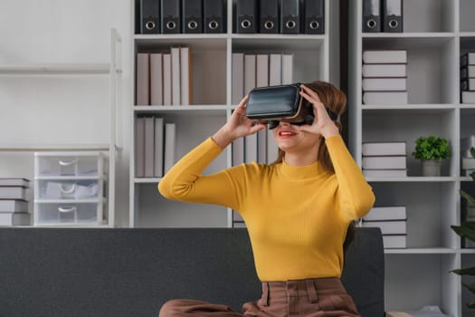 Charming Asian woman plays games in virtual reality through VR glasses on the sofa with a fun and entertaining time in your living room at home.