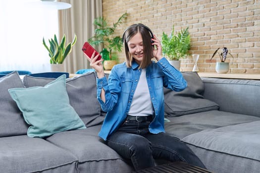 Young happy attractive cheerful woman in headphones listening to music, sitting on sofa at home, in living room, singing along with her eyes closed. Rest, lifestyle, fun, joy, good mood, youth concept