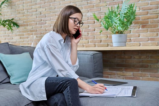 Young female university student studying at home sitting on sofa, living room, talking on cell phone, writing in notebook. Education knowledge youth applications Internet online services for learning