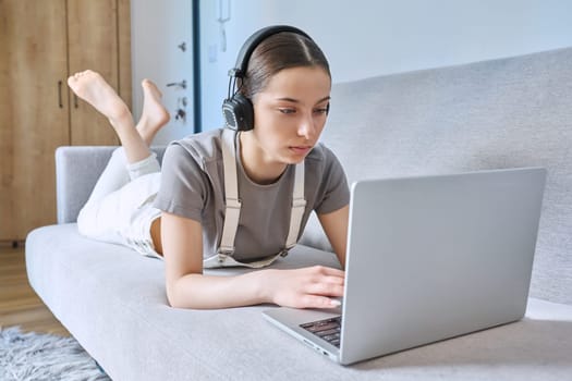 Teen girl in headphones at home on couch, looking at laptop webcam, typing having video chat call conference. Technology for learning leisure study communication modern lifestyle adolescence concept