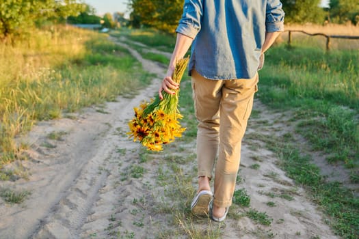 Back view, woman walking on the road with a bouquet of yellow flowers, summer nature, sunset light, countryside