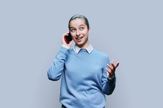 Joyful emotional surprised happy cheerful teenage student, girl 15, 16 years old talking on mobile phone on gray studio background. Lifestyle, communication, adolescence concept