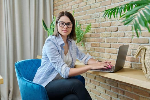 Portrait of young woman sitting at chair with laptop computer, smiling female looking at camera. Business, work, leisure, freelancing, communication concept