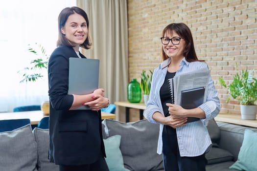 Two happy smiling business women looking at camera standing in office. Successful business, teamwork cooperation collaboration, work job workplace, leadership execution company career people concept