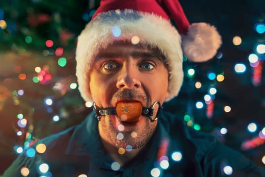 Man with mandarin in his mouth as BDSM sex toys on the Christmas tree background. Concept of fatigue of celebrating New Year's Eve.
