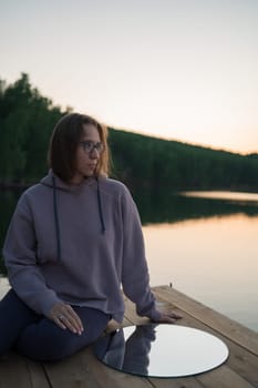 Woman sitting on the pier at lake with circle mirror, summer sunset