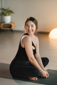 A girl in black sportswear is sitting on a yoga mat and smiling.