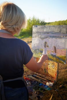 Adult female artist painting picture on the coast of water of a river or lake in nature and trees and greenery in the background