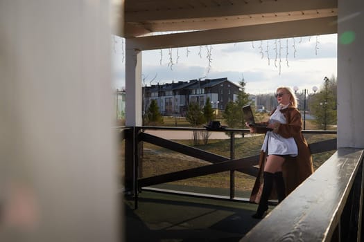 Fashionable beautiful blonde girl is working with laptop in gazebo on sunny autumn, spring day. Businesswoman, entrepreneur, student, manager works outdoors against the backdrop of natural landscape