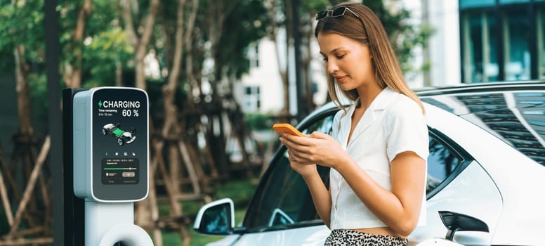 Young woman travel with EV electric car charging in green sustainable city outdoor garden in summer. Urban sustainability lifestyle by green clean rechargeable energy of electric BEV vehicle innards