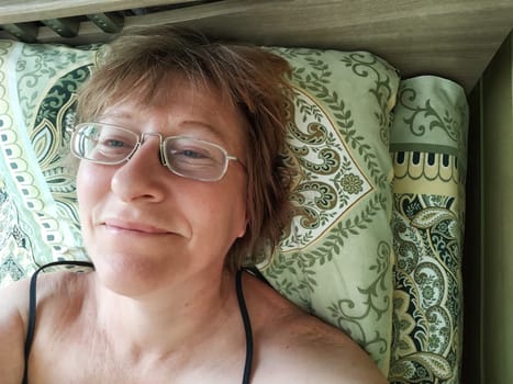 A shaggy, funny middle-aged woman in glasses takes selfie on her smartphone in the bed in early morning. A blogger girl with disheveled hair takes pictures of herself for a blog. Daily routine