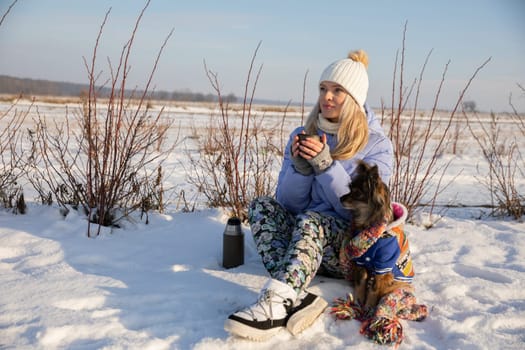 A woman gets lost outside the city. She sits in the snow and tries to warm up by drinking a hot drink from a thermos. Next to its owner sits a small breed dog.