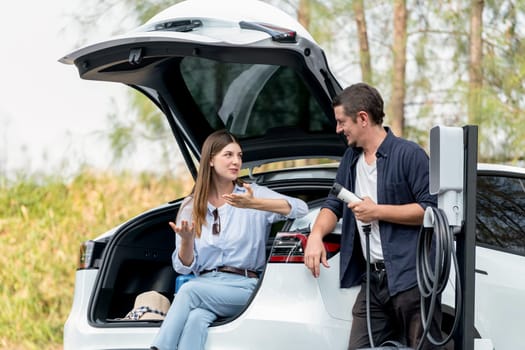 Lovely young couple recharging battery for electric car while relaxing and sitting on the trunk during road trip travel EV car in autumnal forest. Eco friendly travel on vacation during autumn. Exalt