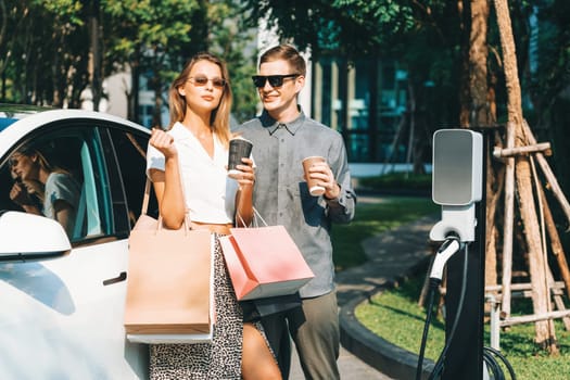 Young couple travel with EV electric car charging in green sustainable city outdoor garden in summer shows urban sustainability lifestyle by green clean rechargeable energy of electric vehicle innards