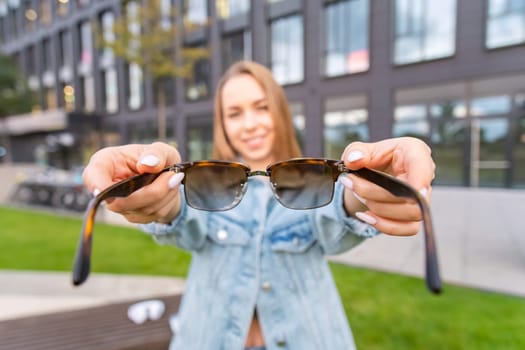 Young blonde woman holding eyeglasses standing near a modern building, view from first person