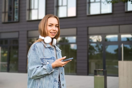 Smiling young woman with headphones on neck looks in camera holding phone. Joyful lady in denim jacket texts best friend after work to hang out
