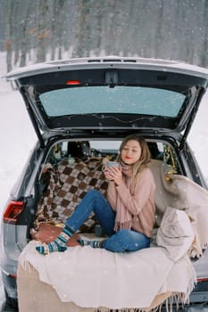 Young woman sits with a mug of coffee squinting in pleasure in the trunk of a car in a winter forest. High quality photo