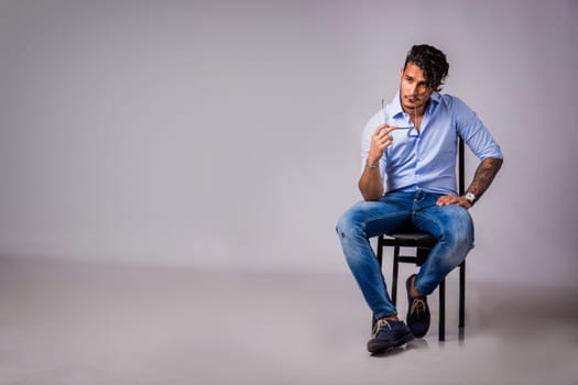 A young handsome man sitting on a chair holding his sunglasses. Photo of a man thinking while sitting on a chair in studio shot on neutral grey background