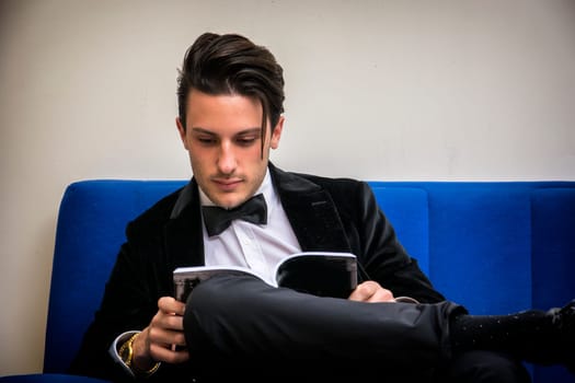 A man in a tuxedo sitting on a blue couch reading a book