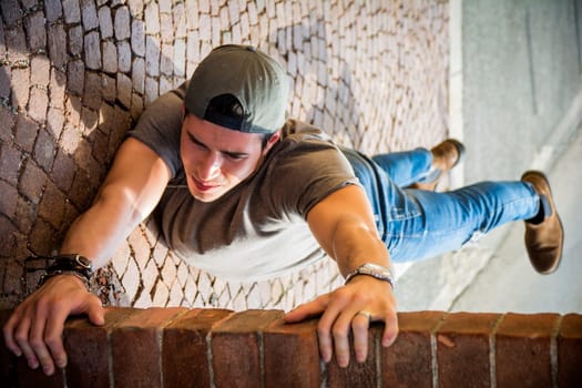 A young man hanging from a brick wall. Optical illusion shot