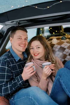 Smiling woman leaning against man sitting with mugs of coffee in car trunk. High quality photo