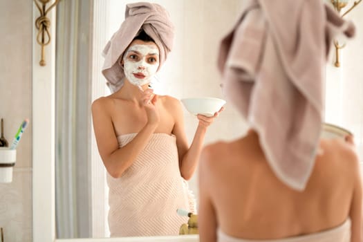 A young girl in a towel on her head and body stands near the mirror in the bathroom and applies a clay mask to her face, the woman takes care of her health and beauty.