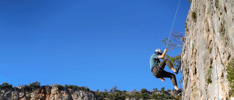 A young man is engaged in rock climbing, mountaineering in a beautiful mountainous area with red rocks, an athlete in a helmet descends from the top against the blue sky on a rope.
