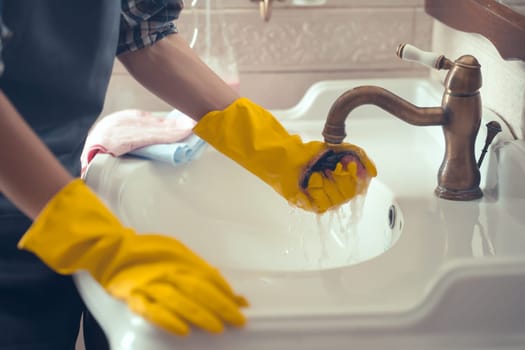 A young girl in an apron and yellow rubber gloves on her hands is cleaning the bathroom, washing the sink with detergent and disinfectant. The woman creates cleanliness at home.