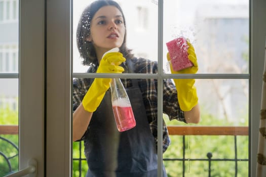 A young girl in yellow rubber gloves and a uniform washes the windows thoroughly with a detergent, wiping them dry with a rag, the woman is cleaning the house.