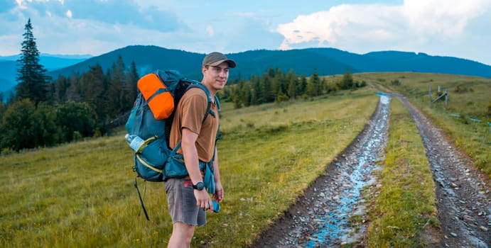 A young man with travel equipment and a large backpack goes on a trip, walks along the road to the national park for hiking in the wild, among the mountains and coniferous forests on a sunny day.
