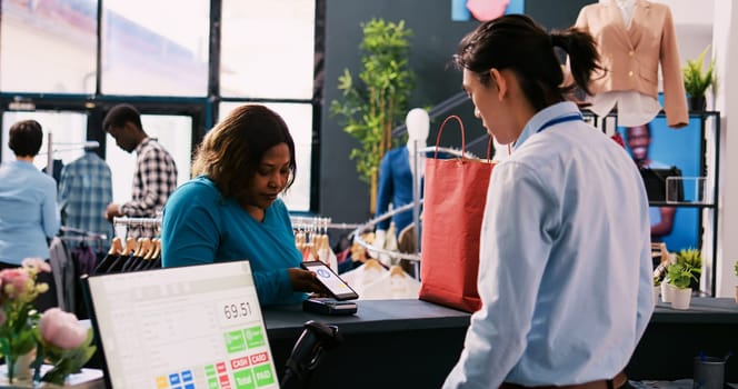 Woman paying with smartphone at pos terminal at counter desk, buying fashionable clothes in clothing store. African american client shopping for formal wear in modern boutique. Fashion concept