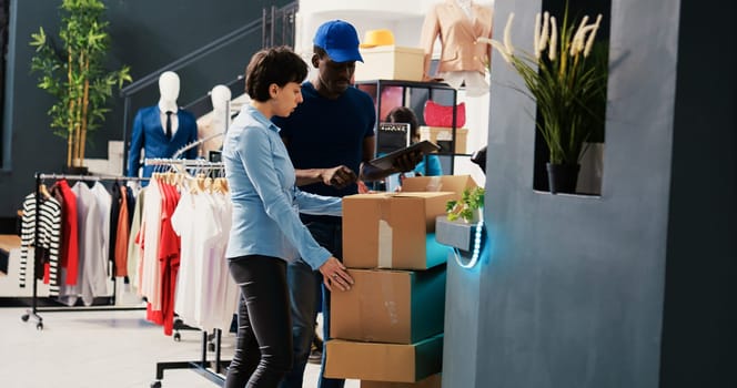 Store workers looking at carton boxes, analyzing distribution report on tablet computer in shopping mall. Manager working at online customers orders, preparing packages for delivery in modern boutique