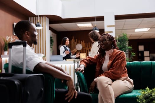 Young couple waiting to do check in at hotel reception, relaxing on couch in luxury lounge area. African american people sitting in modern resort lobby before filling in registration forms.