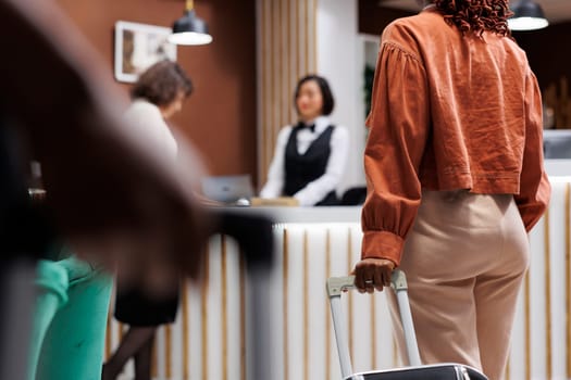 Woman waiting to do check in at hotel, carrying luggage at reception area with front desk. Young female guest preparing to register for room accommodation at luxury resort, lobby space.